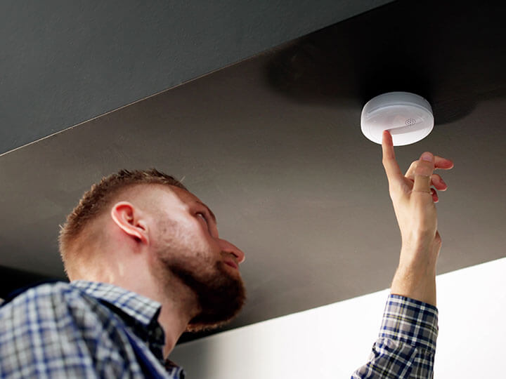 Adult male with beard in a plaid shirt installing a smoke detector and testing it