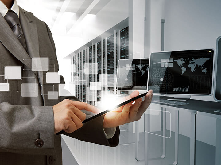 person in grey suit and tie touching a tablet in front of a computer and servers