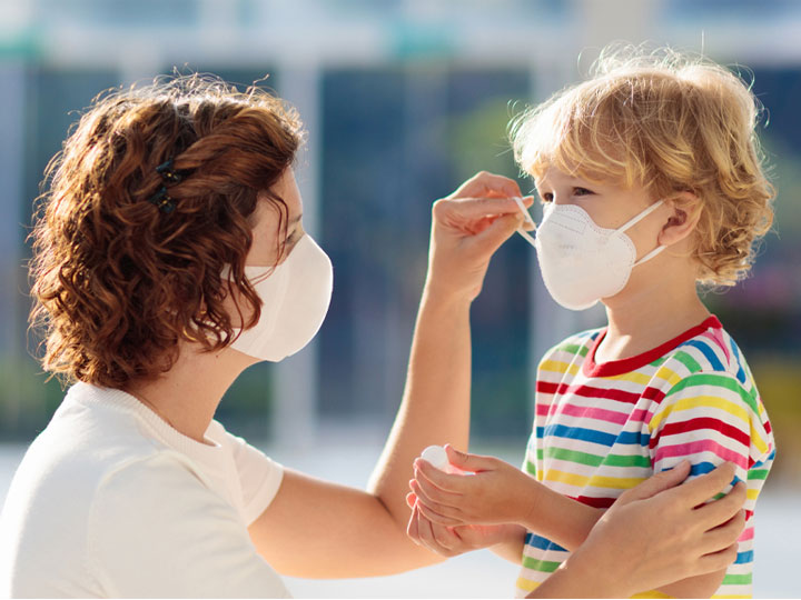 Woman in disposable face bask kneeling to assist a young child with their face mask