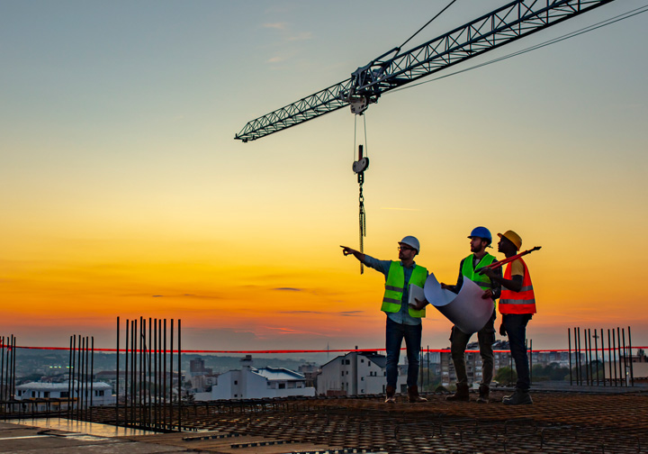 Three people stand on top of a building wearing safety gear, with a crane and an orange sky in the background. 