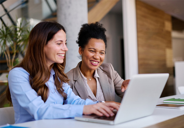 Two women sit in front of a laptop smiling. 