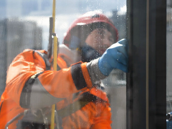 I:nside view of window washer in an orange jumpsuit and safety gear washing windows from dirt and dust