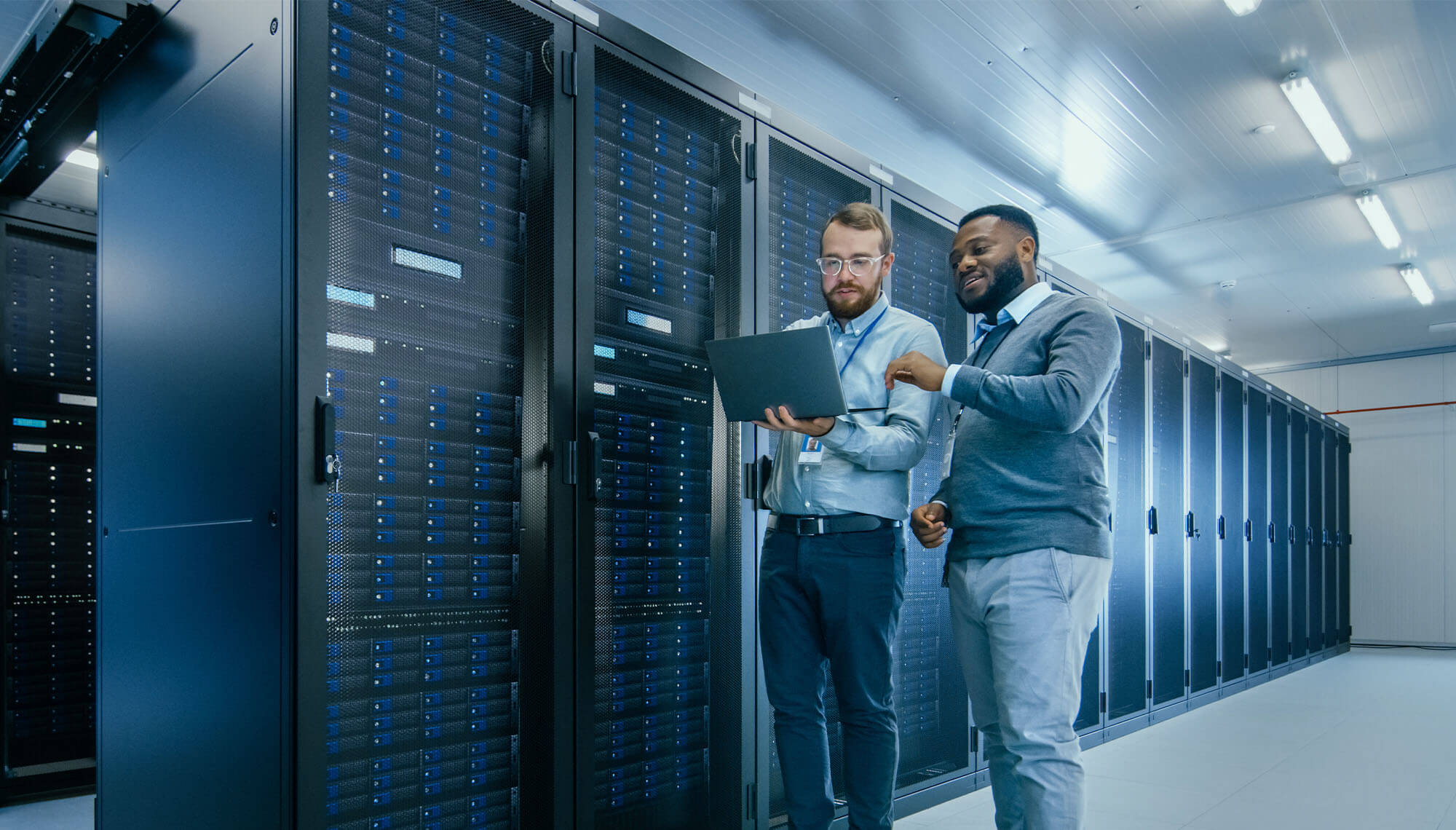 Two men looking at a laptop in a room full of computer servers