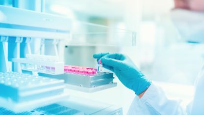 A scientist in a lab coat and gloves places a vial into a tray of pink-tinted samples as part of cosmetics safety testing studies for prohibited or restricted ingredients testing