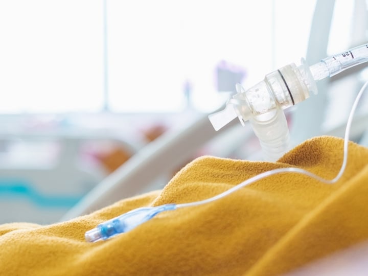 partial view of upper part of a patient lying in a hospital bed covered with a yellow blanket with tubing from a breathing assistance device just showing on the right hand side