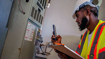 African American Man in His Twenties Photographing an Air Conditioning Unit in the Garage of a Residential Home Wearing Protective Workwear and a Hard Hat
