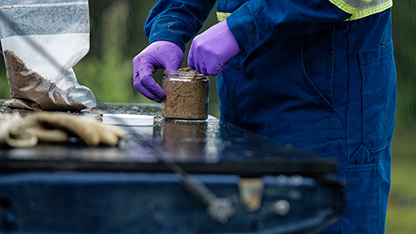 A man in blue coveralls and purple gloves pours a soil-like substance into a jar, focused on his task.